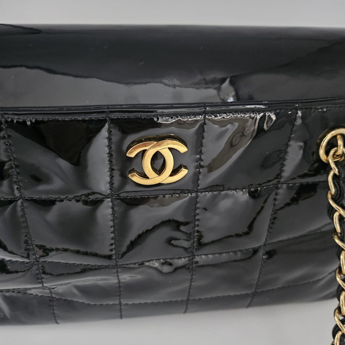 A close-up of a black quilted patent leather handbag with a gold-tone interlocking CC logo on the front, set against a neutral gray background.