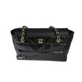 A black quilted patent leather handbag with gold-tone chain straps and a gold interlocking CC logo on the front, photographed against a light gray studio backdrop.