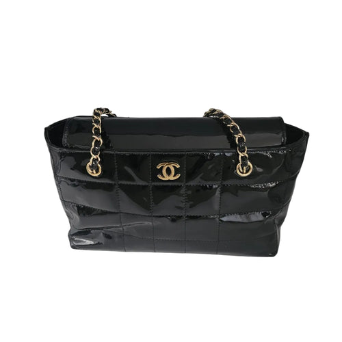 A black quilted patent leather handbag with gold-tone chain straps and a gold interlocking CC logo on the front, photographed against a light gray studio backdrop.