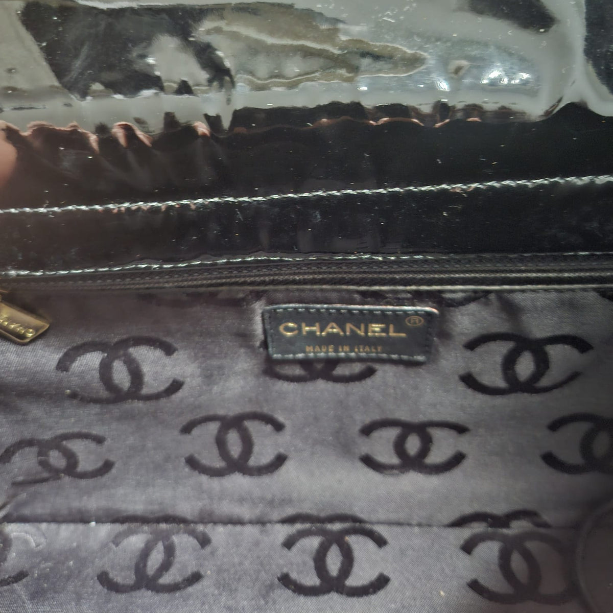 Close-up of a Chanel handbag interior showing a black and gray patterned fabric lining with a gold-tone metal logo plaque reading CHANEL, plus a glossy black zipper track.