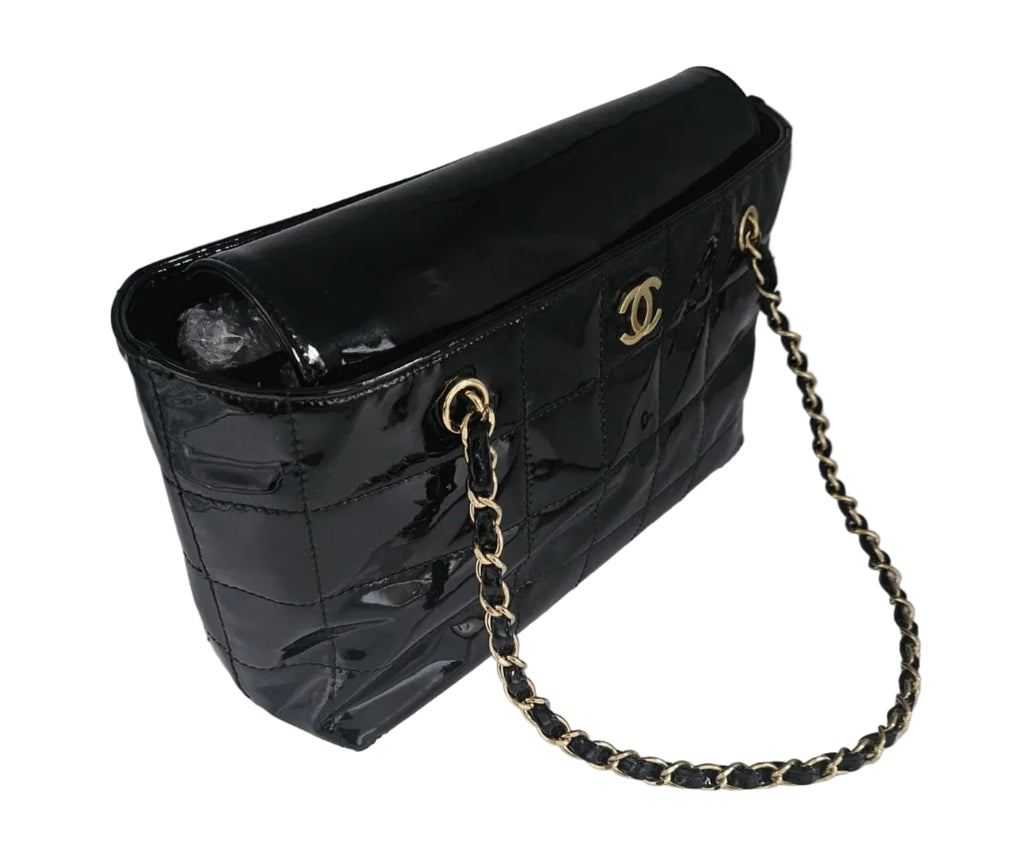 A black patent leather shoulder bag with a quilted front, gold-tone interlocking CC logo, and a gold-and-black chain strap, photographed on a light gray surface.
