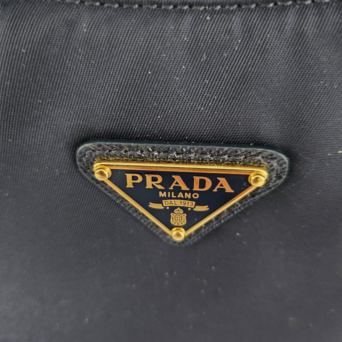 Close-up of a Prada nylon bag with a gold-tone triangular metal logo plaque on a black fabric surface.