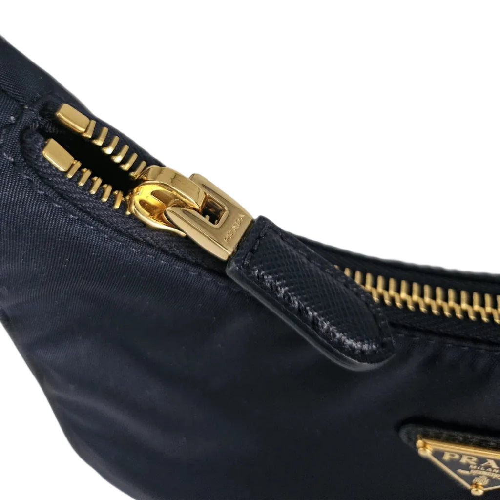 Close-up of a navy blue Prada nylon bag with gold-tone hardware, showing a zipper pull and a small triangular Prada logo plaque.