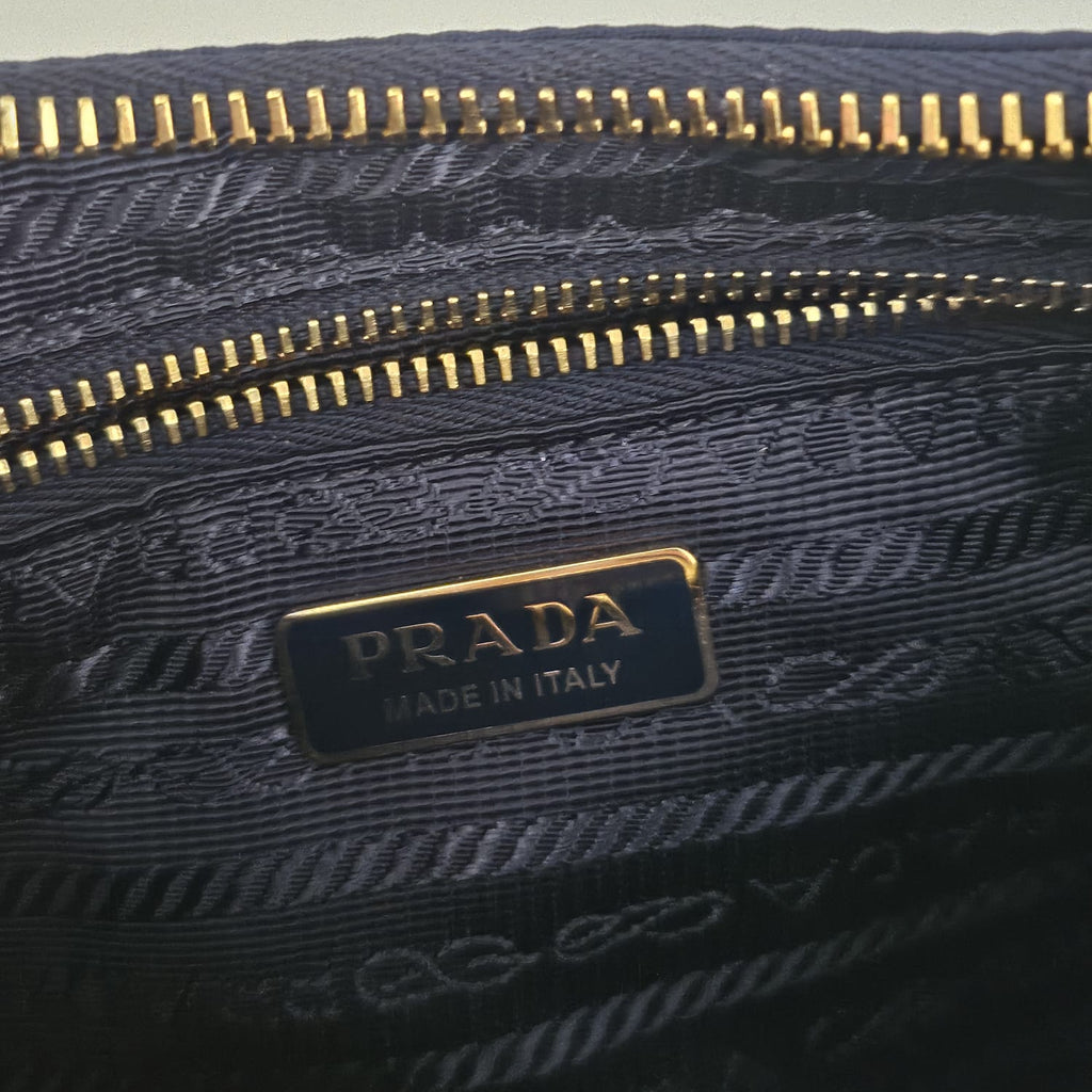Close-up of a Prada bag featuring a dark navy blue jacquard fabric with gold zippers and a gold-tone metal logo plaque that reads 'PRADA MADE IN ITALY'.